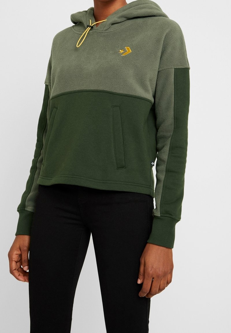 Personne portant un sweat à capuche polaire bicolore vert avec poches avant, un cordon jaune et un pantalon noir sur un fond uni.