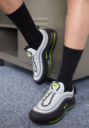 Person trägt schwarze Socken und graue, schwarze und neongrüne Nike-Sneaker, steht auf blauem Teppich in der Nähe eines grauen Spinds.