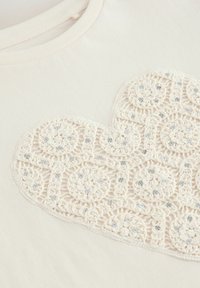 Cremfarbener T-Shirt mit einem strukturierten, gehäkelten Herzdesign, verziert mit hellblauen Pailletten, runder Form und weichem Stoffgefühl.