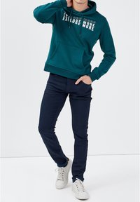 Felpa con cappuccio color teal con stampa "EXPLORE MORE", tasca frontale, abbinata a jeans slim-fit blu scuro e sneakers bianche.