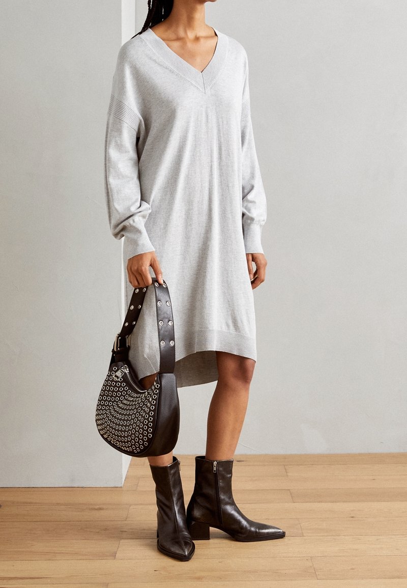 Femme portant une robe ample gris clair à col en V et manches longues, tenant un sac à main noir clouté, et portant des bottines noires à talons carrés.