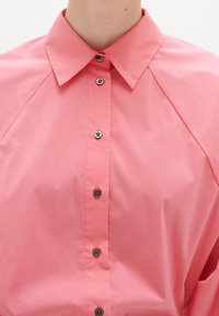 Camicia button-up rosa con colletto classico, caratterizzata da bottoni neri, texture liscia e maniche raglan per una vestibilità rilassata.