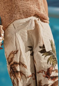 Pantalons larges de couleur crème avec un motif texturé, dotés d'une taille à cordon et d'un imprimé tropical de feuilles et d'oiseaux dans des tons de marron.