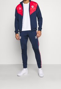 Muž v tmavě modré a červené Adidas Arsenal teplákové soupravě s bílými teniskami stojící na pozadí světle šedé barvy.