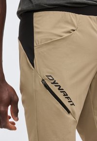 Κοντινό πλάνο ενός ατόμου που φοράει μπεζ παντελόνια Dynafit με μαύρο πλευρικό πάνελ, τσεπάκι με φερμουάρ και το λογότυπο «DYNAFIT» στο μηρό.