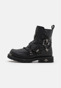 Botines de cuero negro con hardware metálico, múltiples correas, puntera redonda, superficie texturizada y una suela de goma gruesa con un acento decorativo.