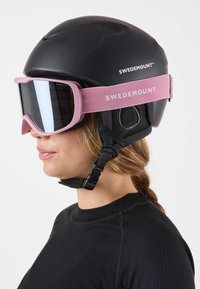 Musta hiihtokypärä, jossa on vaaleanpunainen hihna, jossa lukee "SWEDEMOUNT", sekä suuret peilitut hiihtolasit pehmeällä kehyksellä, jotka ovat päässä olevan henkilön päällä.