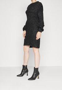 Robe noire en tricot avec une finition texturée, dotée de longues manches bouffantes et d'un col rond, associée à des bottines noires à zip.