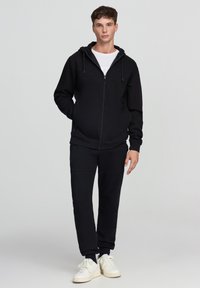 Giovane in felpa nera con zip e pantaloni jogger abbinati, con sneakers bianche, in piedi contro uno sfondo grigio chiaro uniforme.