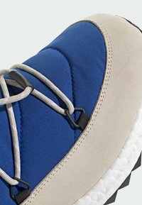 Blå och beige sportskor med en vadderad ovandel, texturerade mockadetaljer, elastiska snören och en vit dämpad sula med svart mönster.