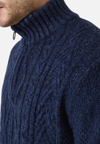 Marineblauer Strickpullover mit hohem geripptem Kragen und Front-Reißverschlussdetail, das einen strukturierten, dicken Stoff und ein auffälliges Muster präsentiert.