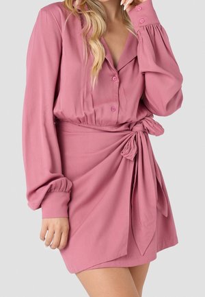 Donna che indossa un vestito rosa a portafoglio con maniche lunghe, bottoni e un fiocco annodato in vita, in piedi davanti a uno sfondo chiaro e semplice.