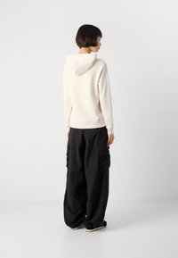 Person with short dark hair wearing a cream hoodie and loose black cargo pants, standing and facing sideways against a plain white background.