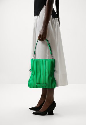 Borsa a pieghe verde con superficie testurizzata, tracolla regolabile e hardware in metallo. Indossata con una gonna bianca e scarpe con zeppa nere.