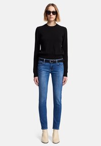 Maglione nero a maglia, jeans slim blu, cintura nera, stivaletti crema alla caviglia e occhiali da sole neri. Tessuto strutturato, design aderente, silhouette moderna.