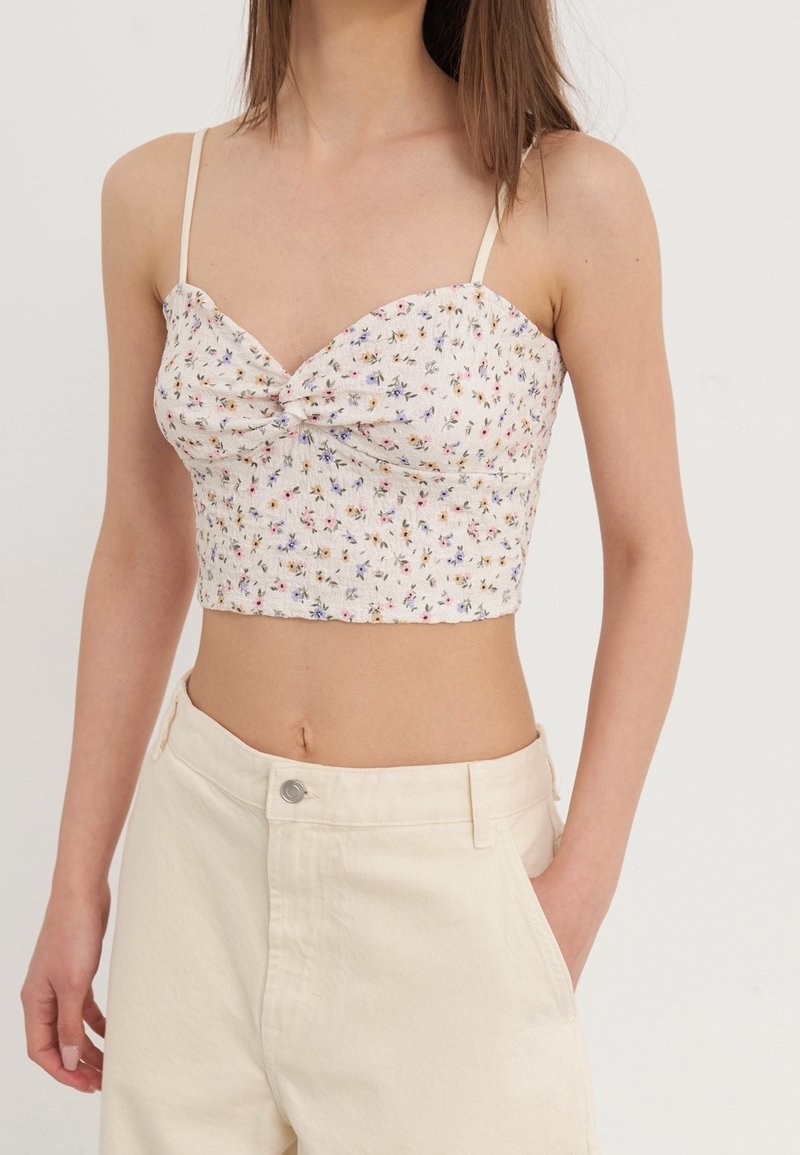 Top court floral en tissu crème, avec un détail avant tordu, des fines bretelles et une coupe ajustée. Associe avec un pantalon taille haute off-white.