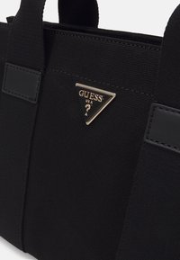 Černá plátěná kabelka s dvojitými tkaninovými rukojetmi, zdobená zlatou trojúhelníkovou kovovou destičkou s nápisem "GUESS USA" a otazníkem.