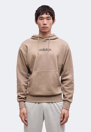Junger Mann trägt einen beigen Adidas-Hoodie mit Fronttasche und grauen Jogginghosen und steht frontal vor einem einfarbigen Hintergrund.