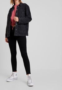 Veste en denim noir avec boutons-pression argentés, deux poches avant, associée à un sweat à capuche rouge et un jean skinny noir. Portée avec des baskets montantes blanches.