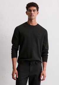 Homme portant un pull noir à manches longues et un pantalon noir se tenant contre un mur blanc texturé et uni.