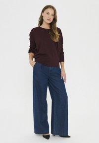 Bordo pleten pulover z dolgimi rokavi v kombinaciji s širokimi temnimi jeans hlačami. Manekenka stoji s rokami v žepih, obuta v črne špičaste čevlje.