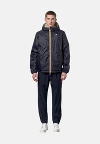 Jeune homme debout, portant une veste zippée à capuche noire, un pantalon noir et des chaussures blanches à lacets, face à l'avant sur un fond uni.