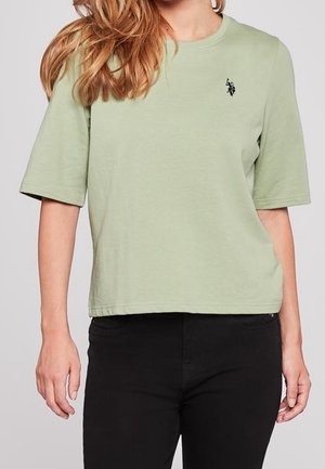 Femme portant un T-shirt à manches courtes vert clair avec un petit logo noir brodé sur le côté gauche de la poitrine et un pantalon noir.