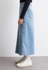 Jeansrock in Hellblau, knielang, mit geradem Schnitt, ausgestattet mit einer Gesäßtasche und sichtbaren Nähten. Kombiniert mit weißen Sneakern und Socken.