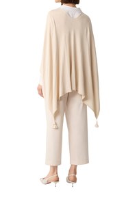 Beige poncho met een zachte textuur, brede snit en franjes aan de zoom, gedragen over een wit shirt en lichtgekleurde broek.