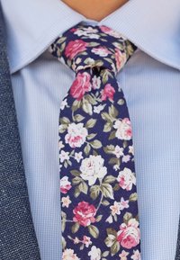 Blomster-slips med marineblå baggrund, prydet med mønstre af lyserøde og hvide roser. Båret over en lyseblå ternet skjorte.