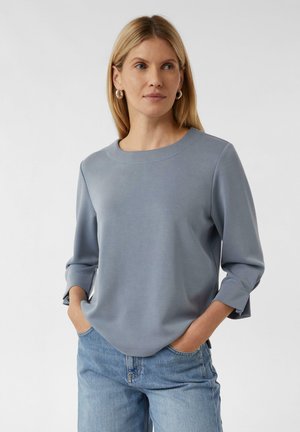 Maglietta blu a maniche lunghe con scollatura rotonda e polsini leggermente svasati, realizzata in tessuto morbido, abbinata a jeans in denim azzurro chiaro.
