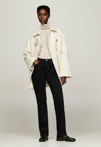 Valge üleüldine mantel kuldsete nuppudega, beež turtleneck, mustad sirged teksad ja mustad loaferid. Sile kangastekstuuri, struktureeritud disain.