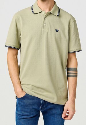Homme portant un polo vert olive clair avec une bordure bleu marine au col et aux manches, un jean bleu, et un tatouage de brassard noir sur l'avant-bras gauche.