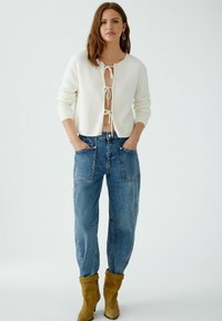 Witte gebreide cardigan met een striksluiting aan de voorkant, gecombineerd met blauwe wijde jeans met grote voorzakken, en beige suède enkellaarsjes.