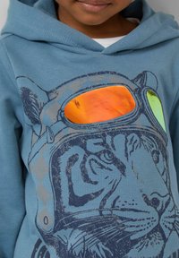 Een kind met een blauwe hoodie met een tijgerillustratie, die reflecterende oranje en groene goggles aan de voorkant draagt.