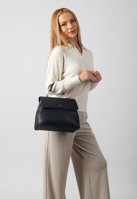 Black handbag with a structured design, gold hardware accents, and a flap closure. Smooth material with a rectangular shape, carried by a woman.