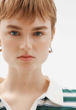 Camisa de cuello con rayas horizontales verdes y blancas. Cabello corto, pendientes de aro y maquillaje neutro. Fondo claro.