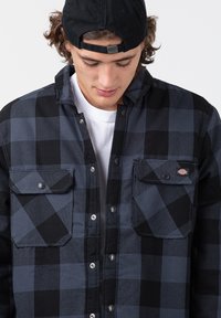 Black and gray checkered flannel shirt with two front pockets, snap buttons, and a tag on the left pocket. Fabric appears textured.
