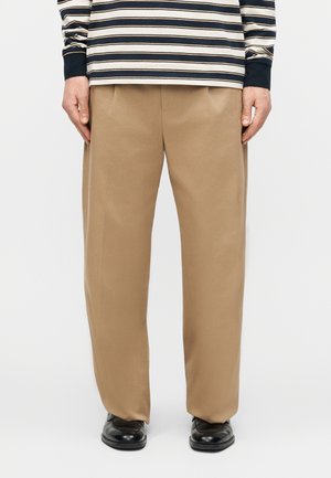 Άτομο που φοράει καφέ παντελόνι φαρδιάς γραμμής, μπλε και λευκή ριγέ μπλούζα με μακριά μανίκια και μαύρα παπούτσια, στέκεται μπροστά σε απλό φόντο.