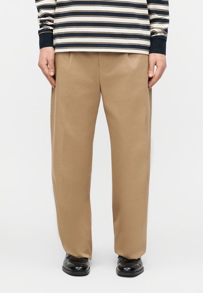 Personne portant un pantalon large couleur sable, une chemise à manches longues rayée marine et blanche, et des chaussures noires, debout devant un fond uni.