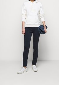 Sweat-shirt à capuche blanche avec une poche frontale et une légère broderie, associé à un pantalon slim fit bleu marine et des baskets blanches, tenant une veste en jean.
