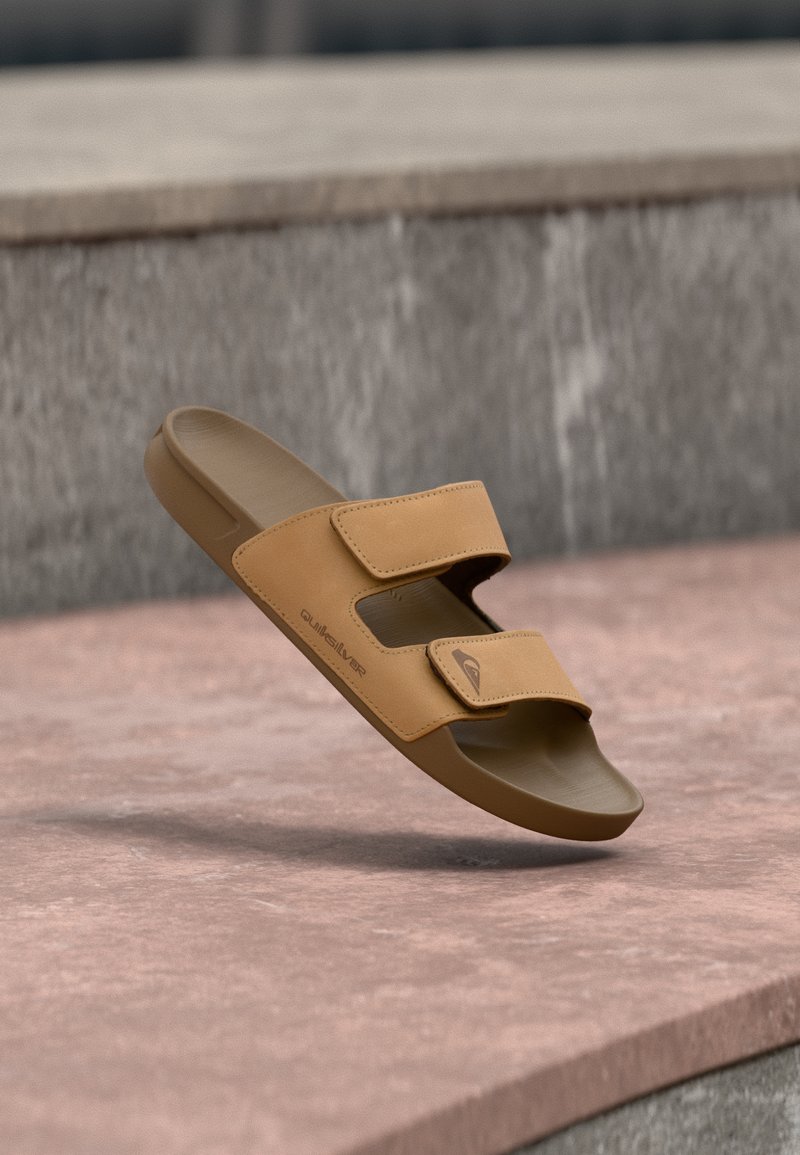 Ροζ σανδάλι slide Quiksilver με δύο ρυθμιζόμενα λουριά, να αιωρείται πάνω από μια επιφάνεια από ροζ και γκρι πέτρα.