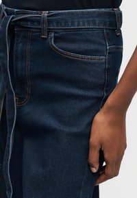 Κοντινό πλάνο του χεριού ενός ατόμου που ακουμπά δίπλα σε σκούρα μπλε τζιν με εμφανή ραφές και δεμένη θηλιά ζώνης.