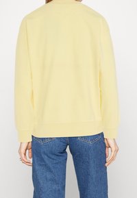 Sudadera amarilla con cuello redondo, mangas largas y ribete acanalado. La tela parece suave y tiene una textura lisa. Combinada con jeans azules.