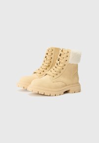 Bottines beiges avec un col en fourrure et des semelles en caoutchouc épaisses, présentant des lacets proéminents et un design robuste, sur un fond uni.