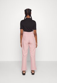 Zwarte top met korte mouwen en hooggepolleerde roze broek, gedragen door een staande persoon met gevlochten haar, met de rug naar de camera op een effen witte achtergrond.