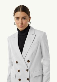 Lichtgrijze dubbelzijdige blazer met bruine knopen, voorzien van een geknipt revers en zijzakken. Draag het over een zwarte coltrui.