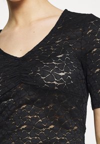 Svart topp i spets, med V-ringning och korta ärmar. Tyget har ett blommigt mönster med texturerade detaljer och genomskinliga partier.