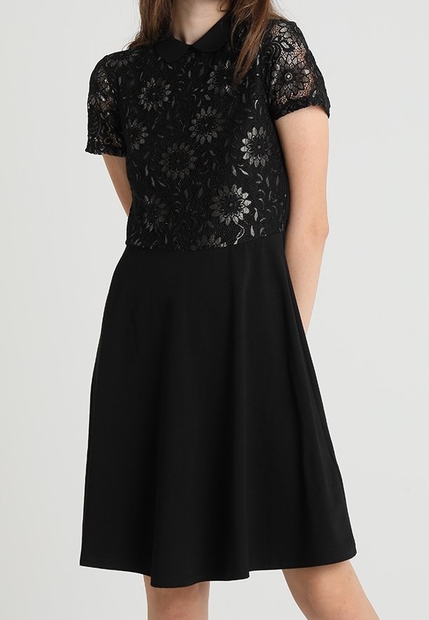 Femme portant une robe noire avec un haut à manches courtes en dentelle florale et une jupe trapèze unie, debout devant un fond clair uni.