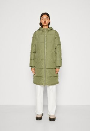 Parka verte rembourrée avec capuche, fermeture éclair et poches latérales. Présente des coutures horizontales et des poignets élastiqués. Portée par-dessus un pantalon blanc.